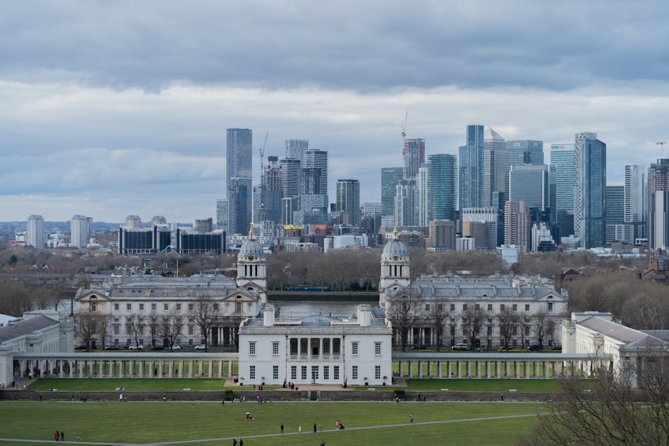 A wide-angle view of the historic Greenwich Royal Naval College, featuring white colonial-style buildings with symmetrical facades, domed towers, and large windows, set against a lush landscaped lawn 